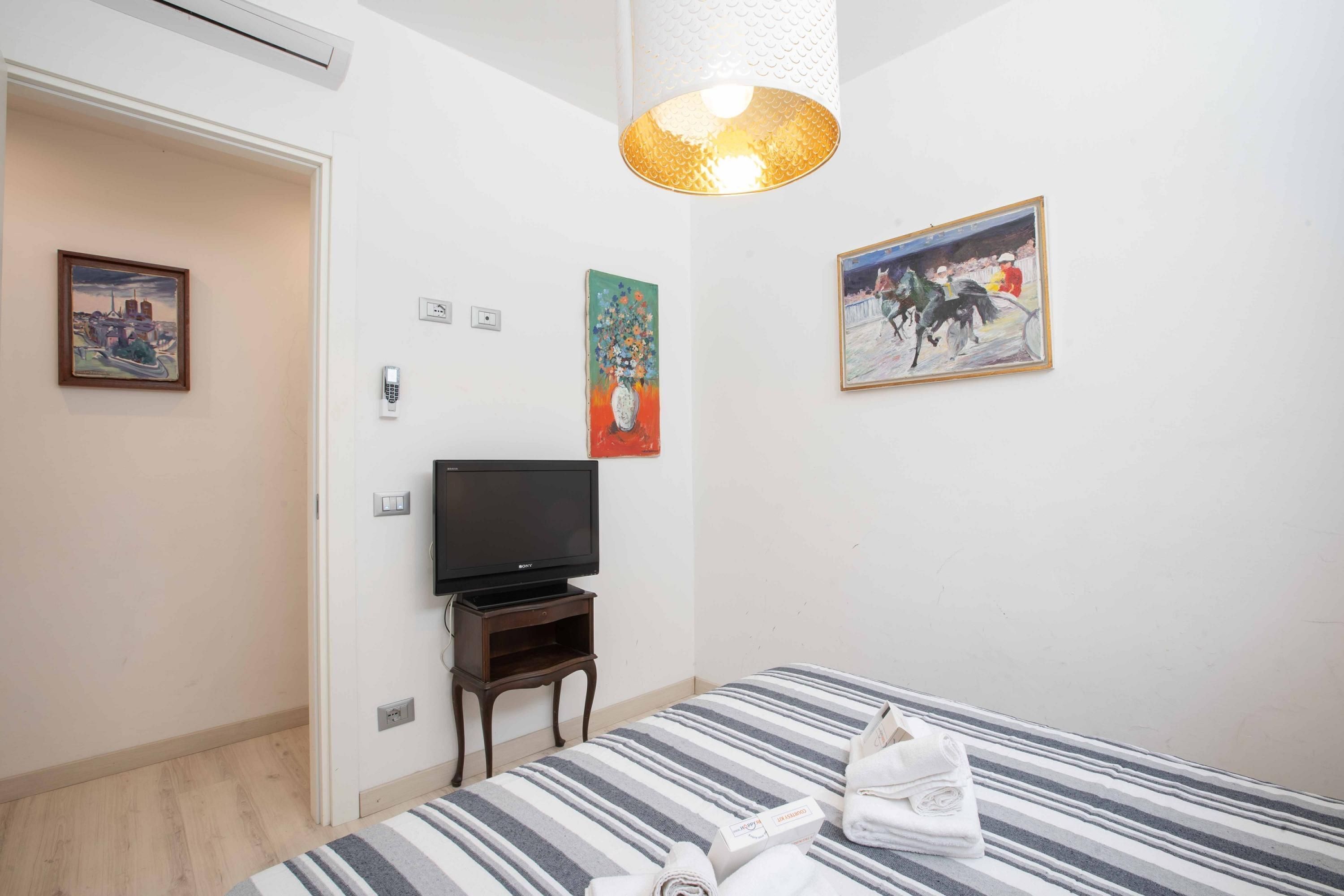 A hotel room with a bed, a television, a painting, and a lamp.