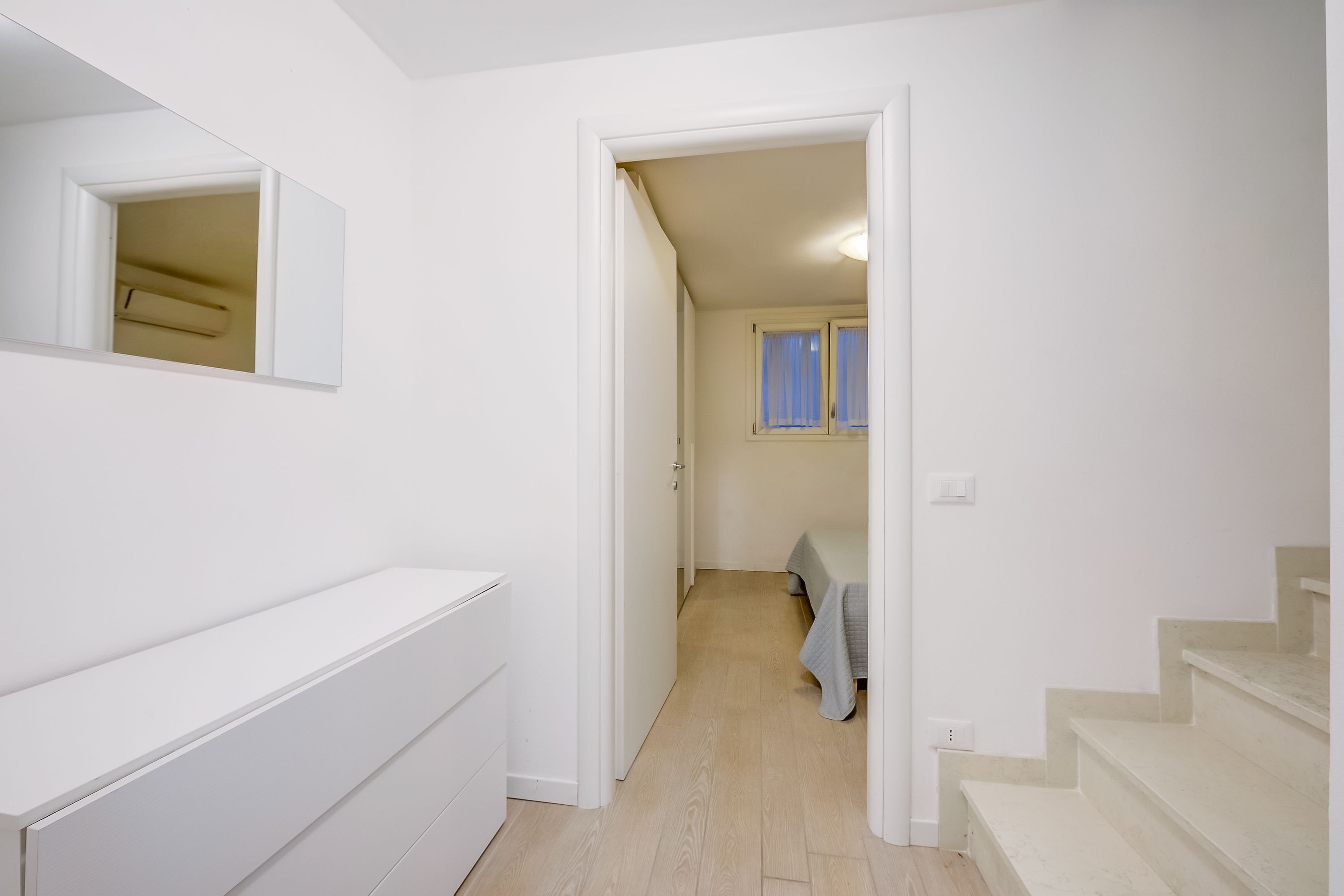 Un pasillo con pared blanca, un espejo, un radiador y una escalera que lleva a otra habitación.