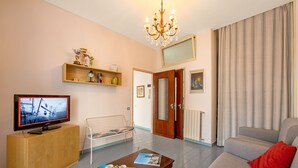 Ein Wohnzimmer mit Fernseher, Sofa und Couchtisch. Eine Tür führt in einen anderen Raum, ein Fenster mit Vorhängen.
