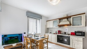 Une cuisine moderne avec des meubles blancs, une table à manger pour quatre personnes, un téléviseur posé sur un meuble en bois et un grille-pain rouge.