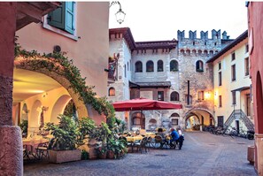 A cobblestone street with outdoor seating, a stone building with arched windows, and a red umbrella. - Casa Elena in Riva del Garda (Riva del Garda)