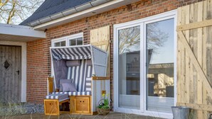 A cozy outdoor seating area with a wooden bench and a striped canopy, adjacent to a brick building with large windows and a wooden door.