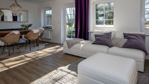 A modern living room with a white sofa, a dining area with wicker chairs, and a large window with purple curtains.