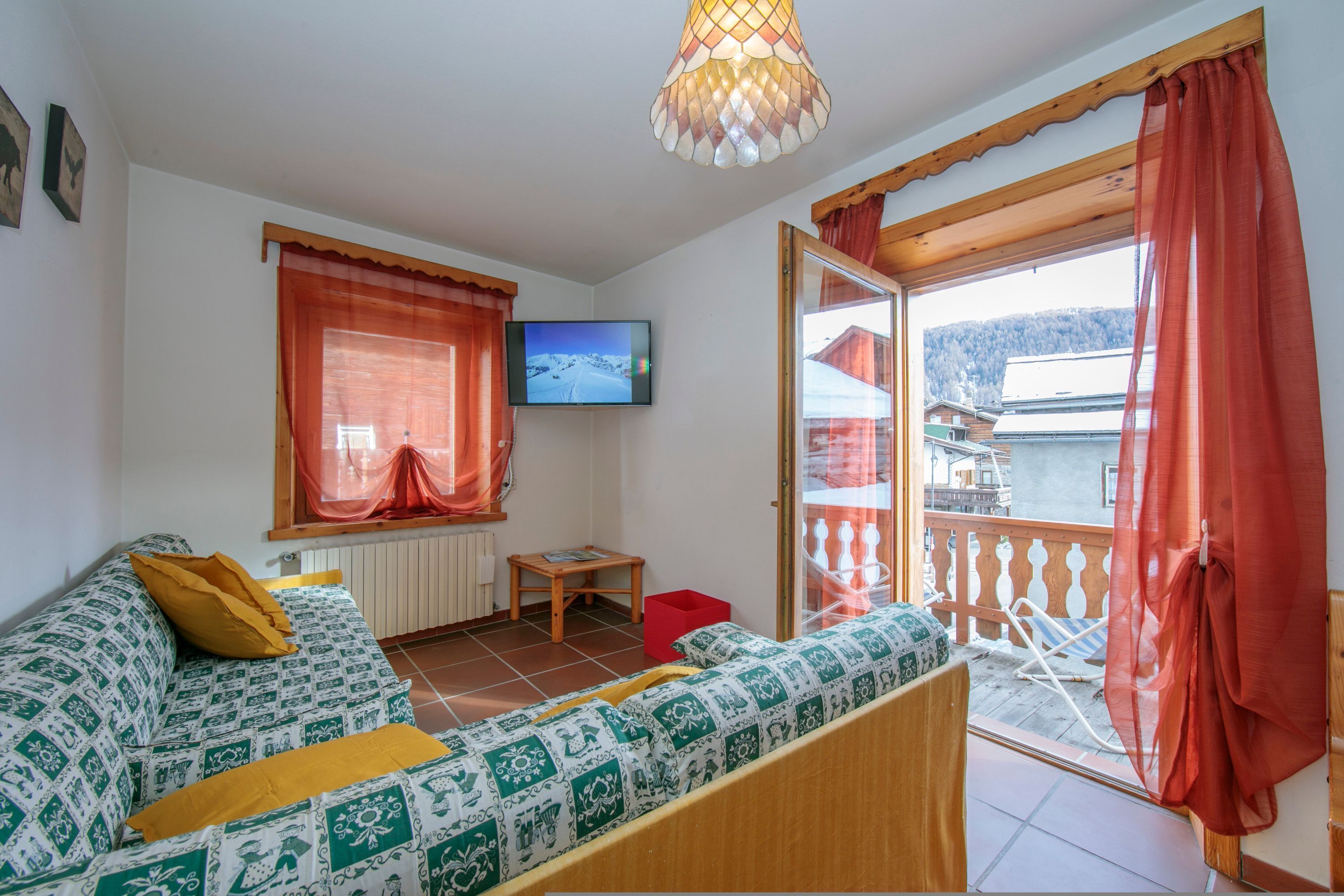 A cozy room with a sofa, a television, and a balcony with a view of a lake and mountains.