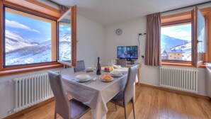 A dining room with a table set for two, a clock on the wall, and a TV displaying a program.
