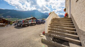 Un edificio con una escalera que lleva a un porche, coches aparcados y montañas al fondo.