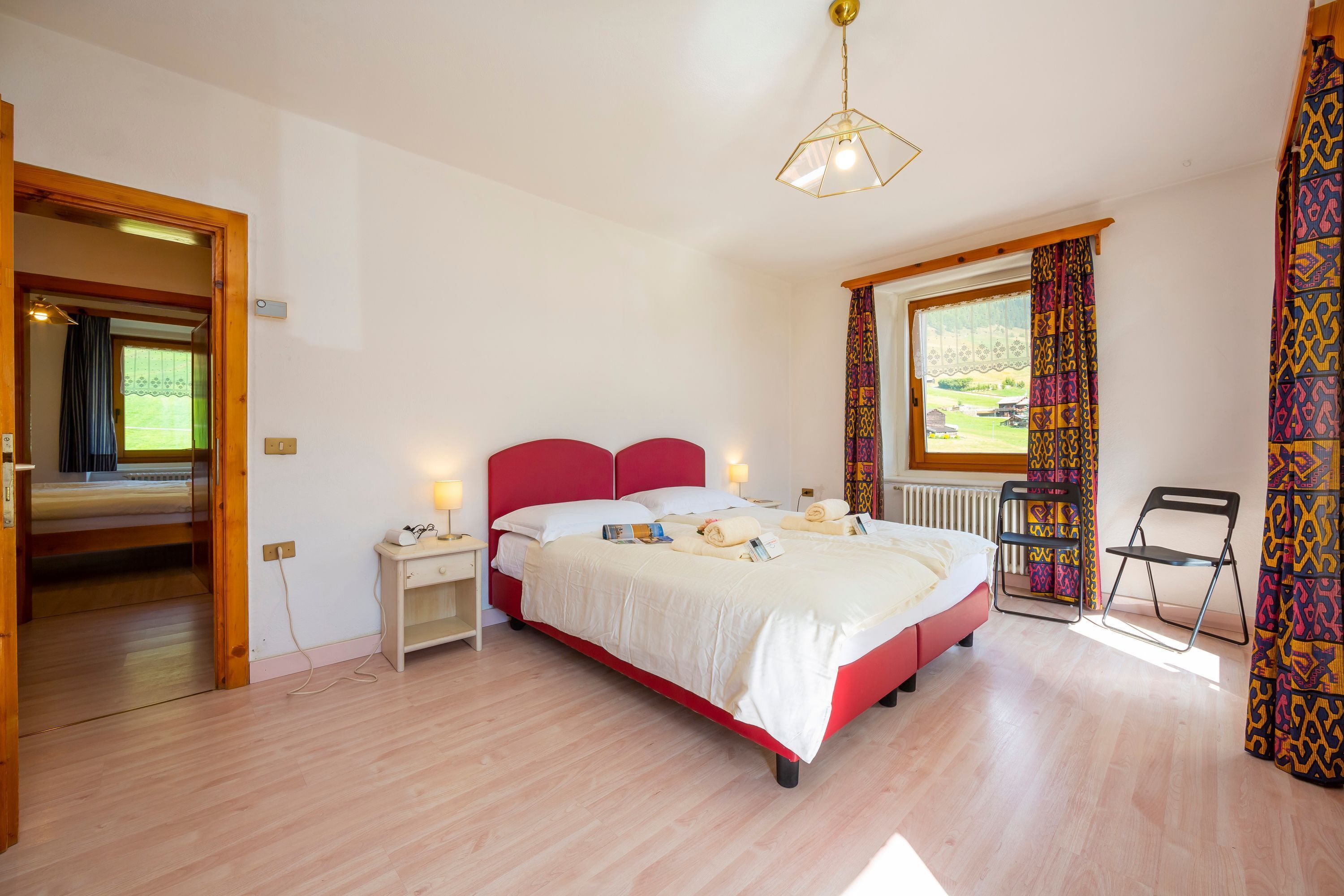 A bedroom with a red bed, white bedding, a bedside table with a lamp, a window with patterned curtains, and a chair by the window.
