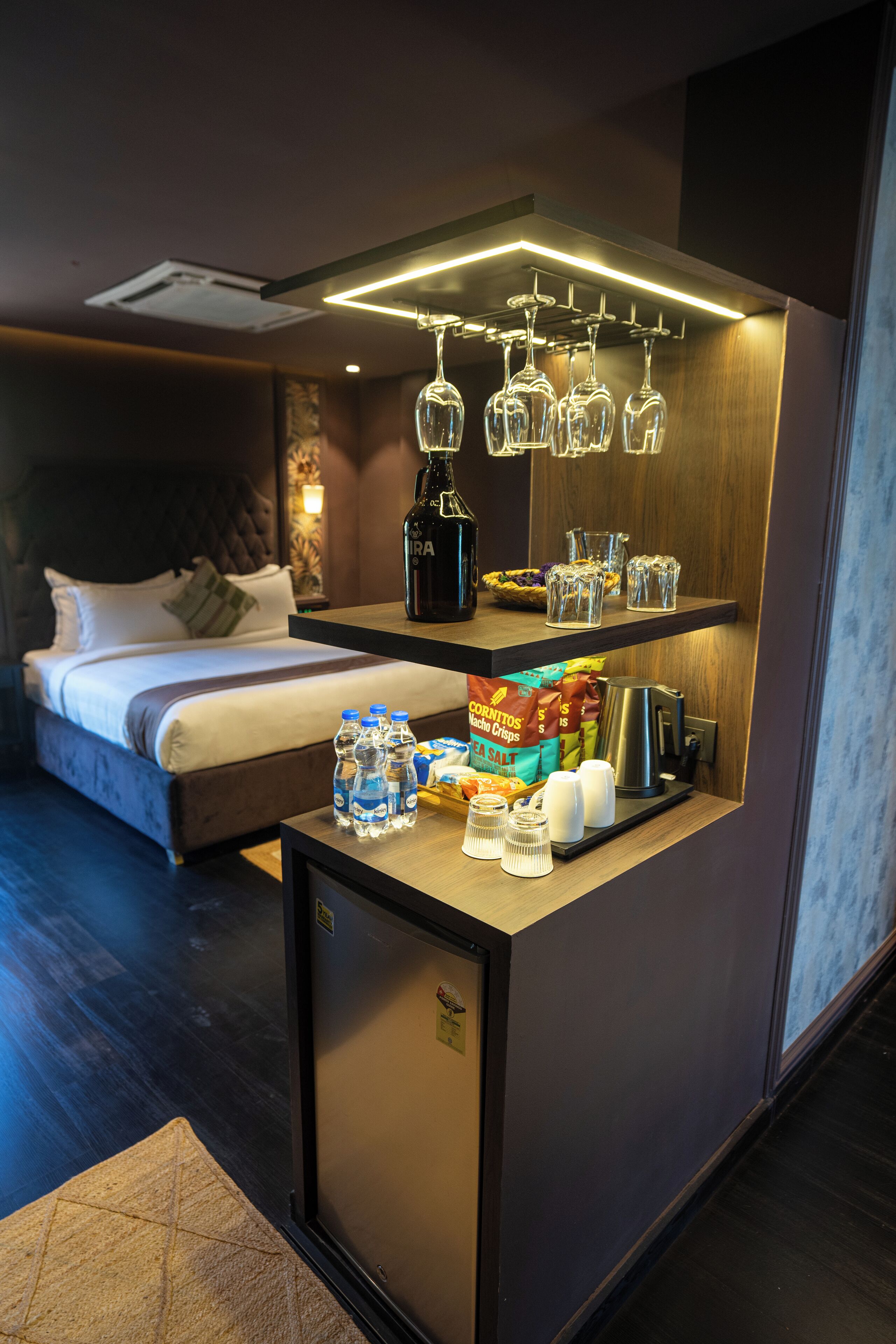 A compact hotel room with a bed, a minibar, and a small bar area with glasses and bottles.
