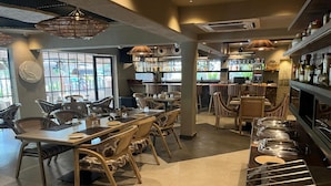 A restaurant with multiple dining tables and chairs, a bar area with shelves of bottles, and large windows allowing natural light. - Origin Boutique Hotel (Arpora)