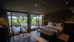 A modern hotel room with a large bed, a seating area with a table and chairs, and large windows offering a view of greenery outside. - Origin Boutique Hotel (Arpora)