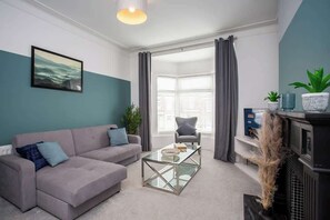 En moderne stue med en grå sofa, et glasbord, en grå lænestol og et stort vindue med gardiner.