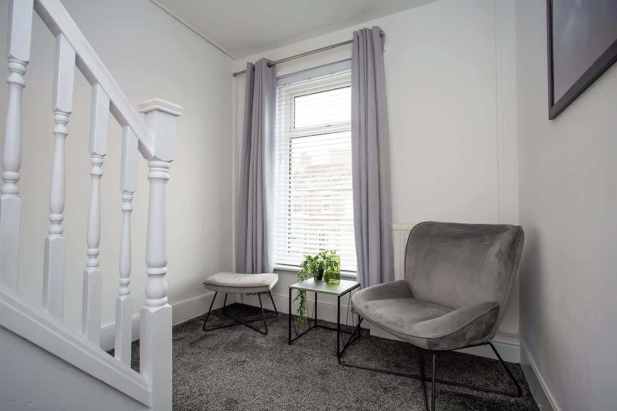 Una habitació amb una escala, una finestra amb cortines, una cadira gris, una taula petita amb una planta i un terra amb moqueta.