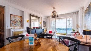 A dining area with a round wooden table, chairs, and a bottle of wine. There is a large window with a view of the sea. - ROMANTIC VILLA VI4412 By Riviera Holiday Homes (Villefranche-sur-Mer)