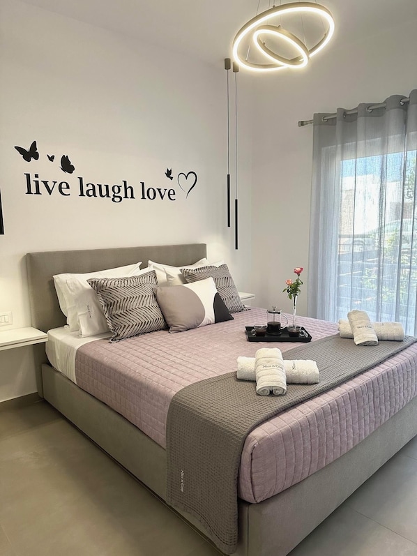 A modern bedroom with a bed, decorative wall art, a bedside table, and a window with curtains. - Nice n Cozy Apartment II in Rhodes Town (C6R8+P53 Rhodes, Greece)