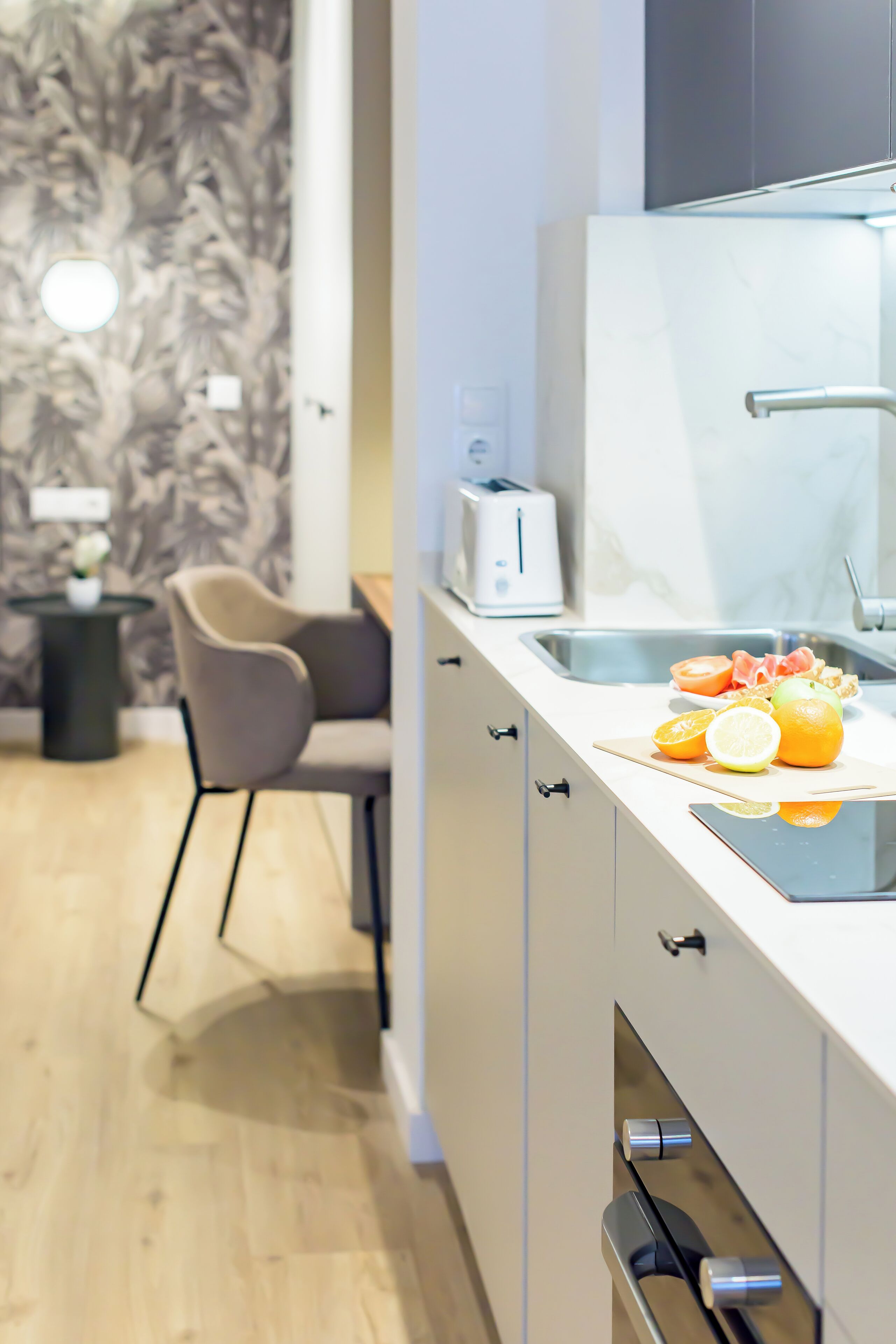 A modern kitchen with a white countertop, a sink, and a toaster. There is a chair and a small table with a vase of flowers in the background.