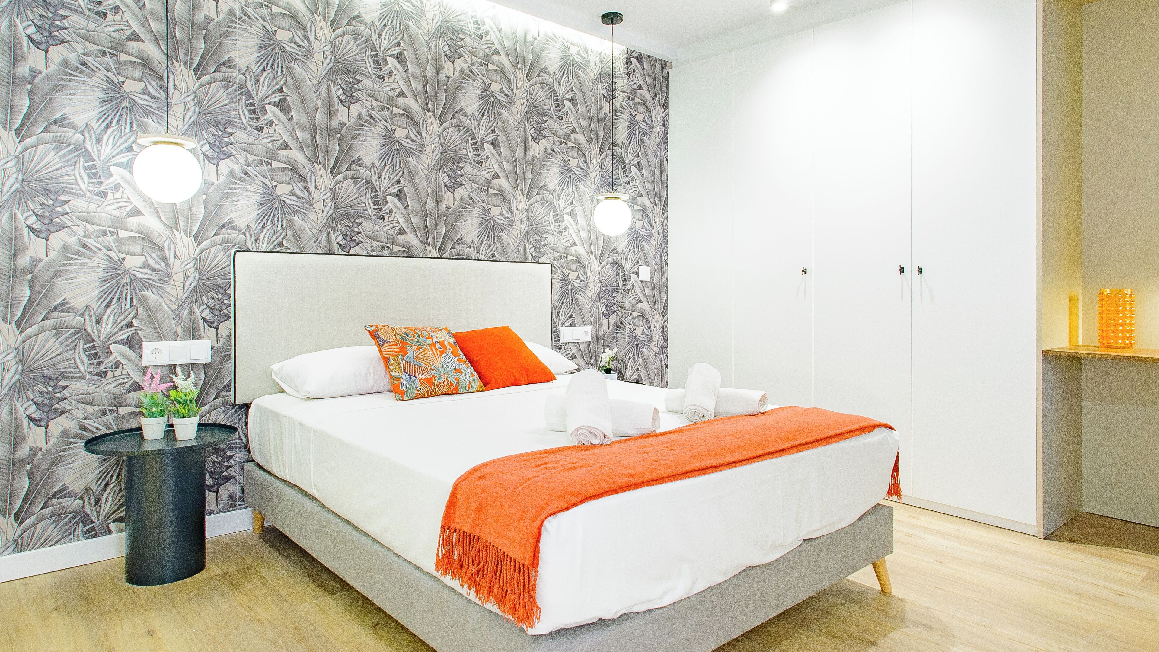 A modern hotel room with a large bed, white bedding, an orange throw blanket, and a tropical leaf-patterned wallpaper.