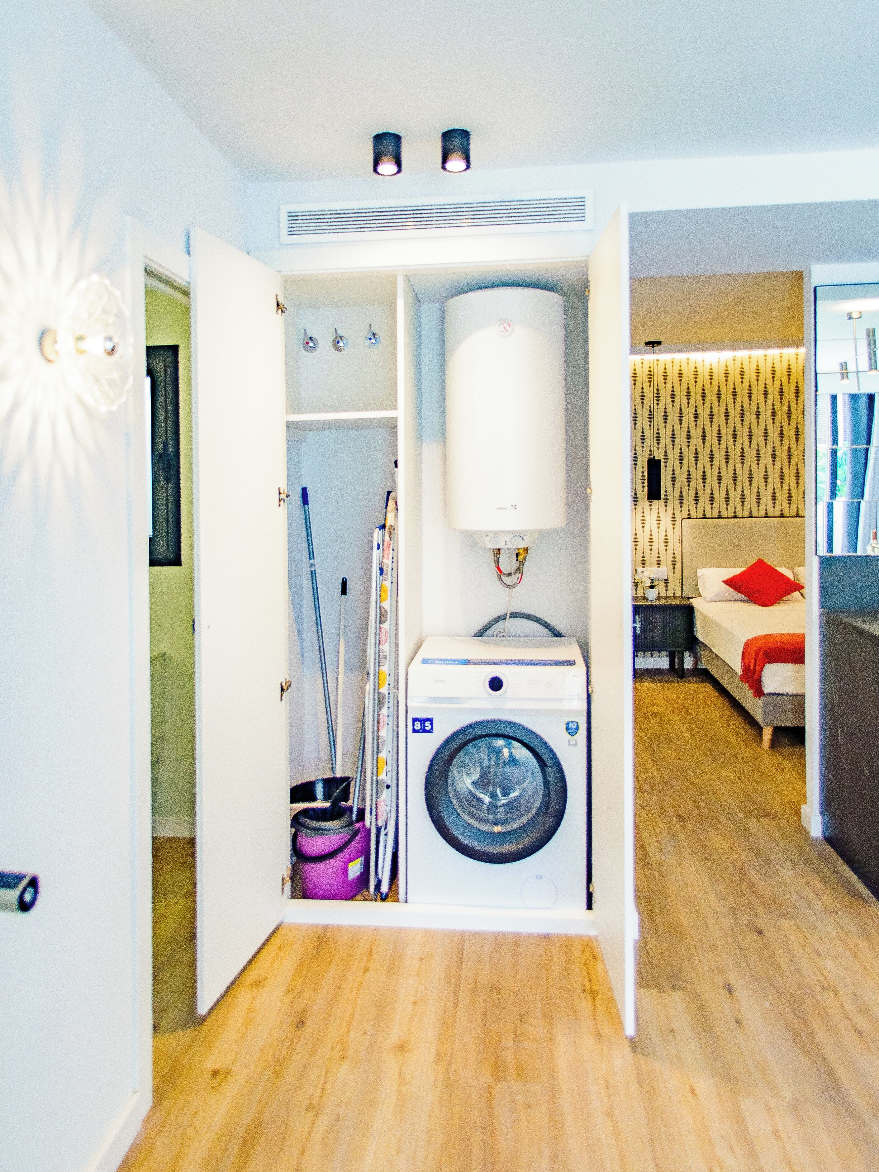 A compact living space with a built-in washer and dryer, a small kitchenette, and a bedroom with a bed and a red pillow.