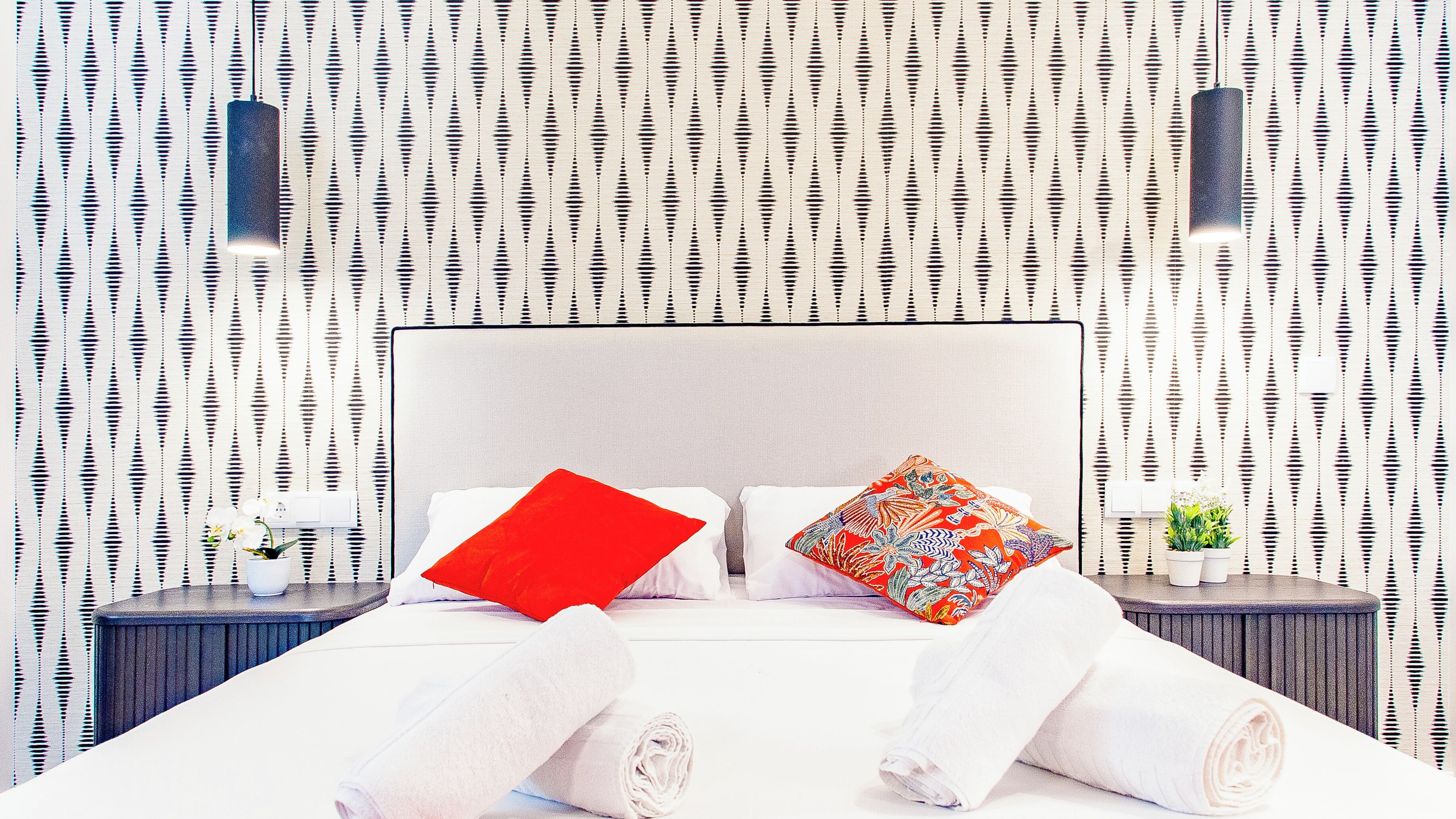 Cama bem arrumada com lençóis brancos, dois travesseiros (um vermelho e um multicolorido) e toalhas enroladas. O quarto tem papel de parede estampado e duas mesas de cabeceira com pequenas plantas em vasos.