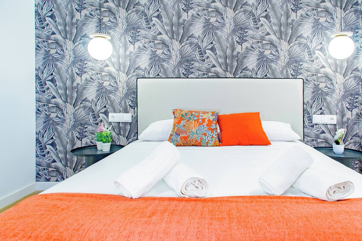 A neatly made bed with white linens and two pillows, one with a colorful pattern and the other solid orange, against a wall with a tropical leaf pattern.