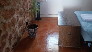Quarto rústico com vigas de madeira aparentes, parede de pedra, pequena janela com cortinas brancas, planta em vaso, cama e cômoda.