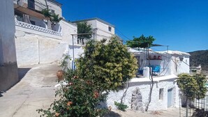 Uma rua estreita e pavimentada com edifícios brancos, uma árvore com flores laranja e um céu azul límpido.