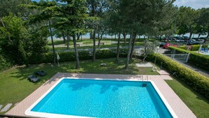 Una piscina rectangular con solárium de baldosas, rodeada de césped, árboles y una vista a un estacionamiento al fondo.