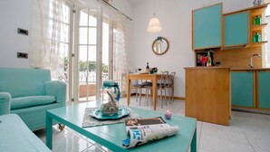 Una sala moderna con un sofá color esmeralda, una mesa de comedor con sillas, una zona de cocina con armarios color esmeralda y un ventanal con cortinas.