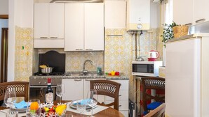Cocina compacta con armarios blancos, un microondas y un fregadero. Hay una mesa de comedor con platos, vasos y una cesta de frutas.