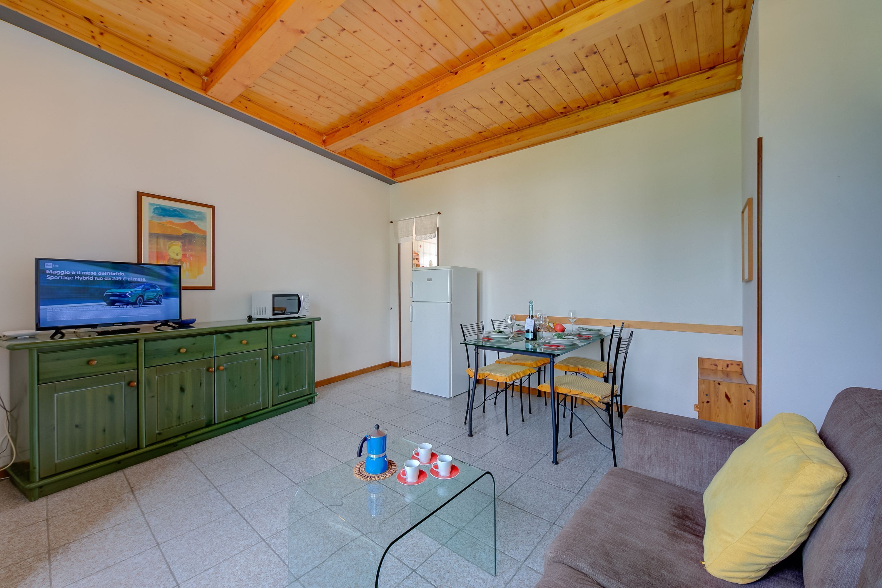 Un salón con techo de madera, un mueble verde, una mesa de centro de vidrio, un sofá y un comedor con sillas y una mesa.