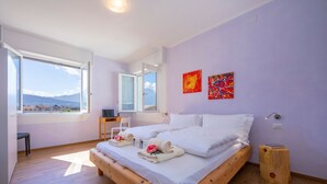 Ein Schlafzimmer mit einem großen Bett, einem Schreibtisch und einem Sessel, ein Fenster mit Meerblick und zwei abstrakte Gemälde an der Wand.