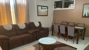 Een woonkamer met een bruine bank, een rond bijzettafeltje en een eethoek met een tafel en stoelen.