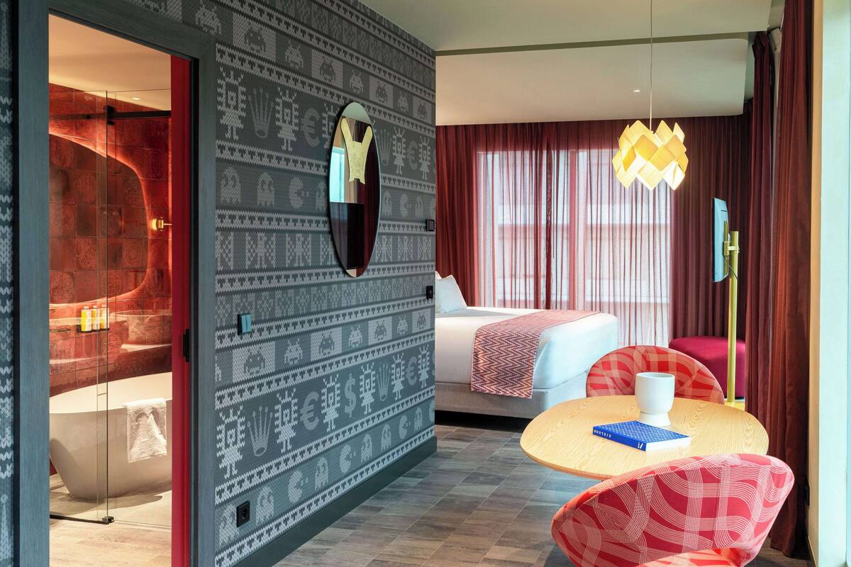 A modern hotel room with a patterned wall, a large mirror, a bed with a red blanket, a round table with a book, and a red and white patterned chair.