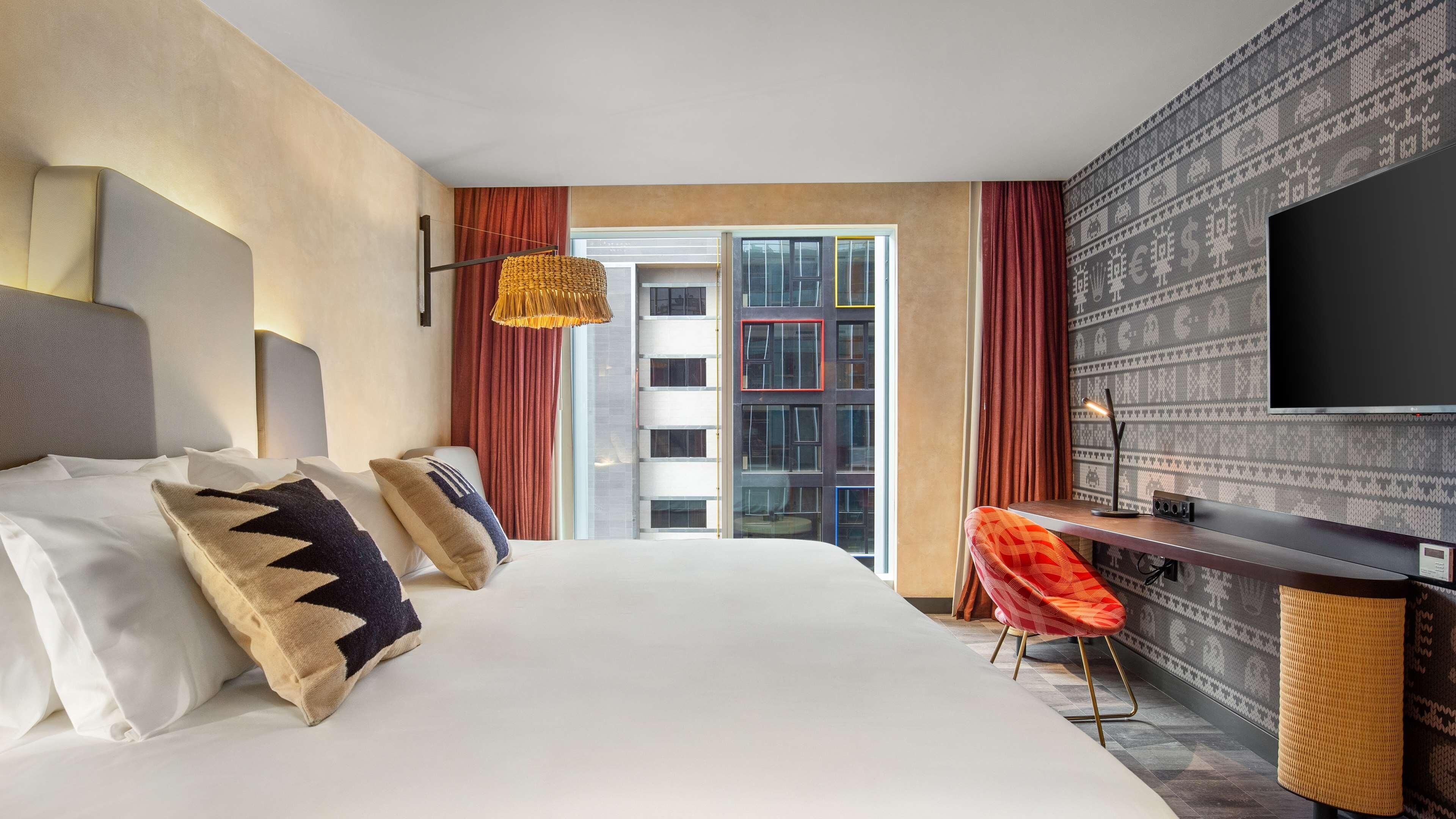 A hotel room with a large bed, a desk, a television, and a view of a building with patterned wallpaper.