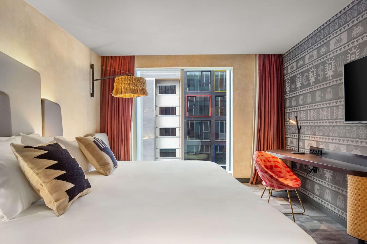 A hotel room with a large bed, a desk, a television, and a view of a building with patterned wallpaper.