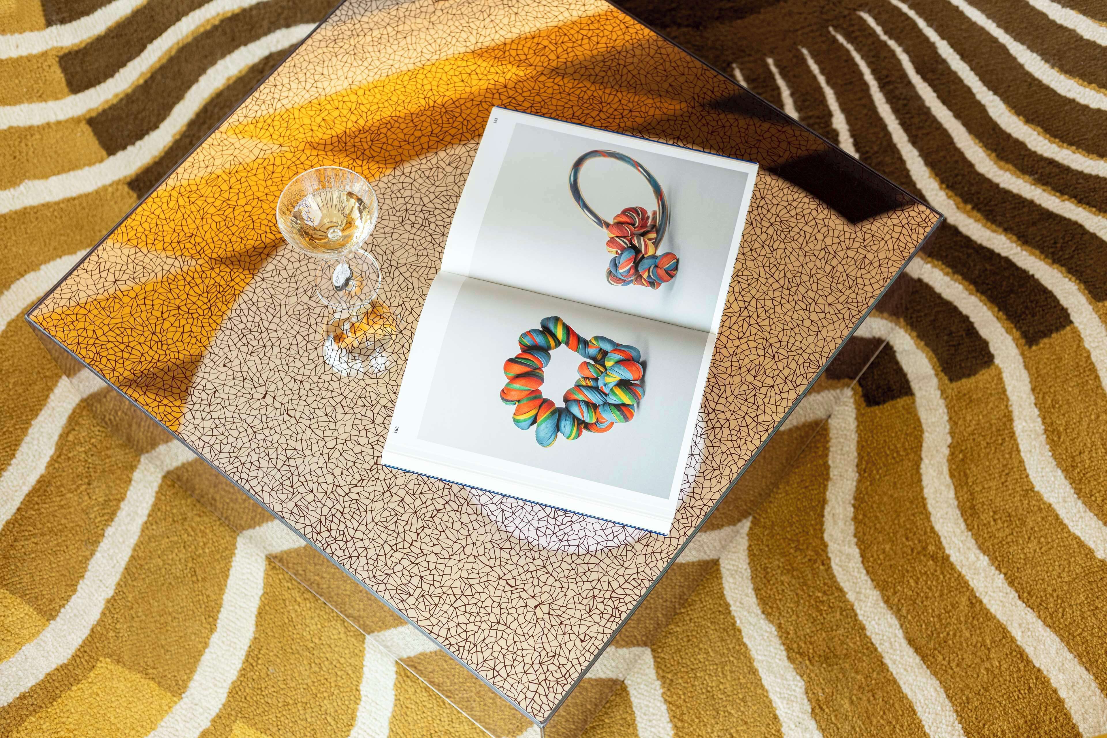 A glass table with a magazine featuring colorful jewelry, a glass of water, and a patterned rug.