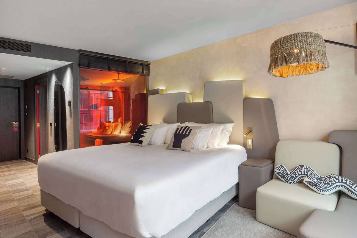 A modern hotel room with a large bed, a bedside table, a chair, and a unique wall-mounted lamp.