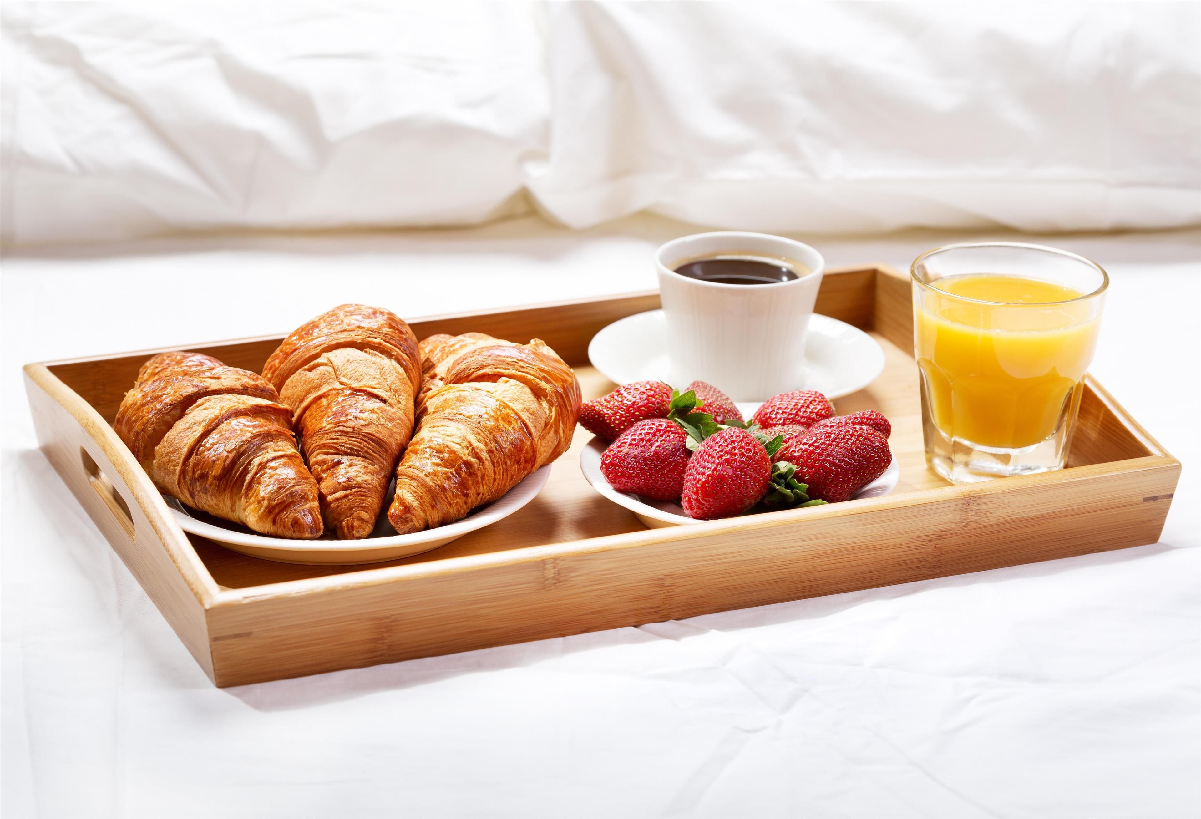 Bandeja de desayuno con cruasanes, fresas, una taza de café y un vaso de jugo de naranja sobre una cama.