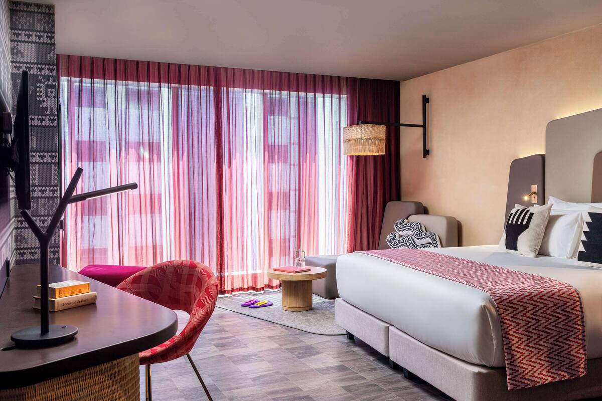 A hotel room with a bed, a desk with a lamp, a chair, a small side table, and a large window with sheer curtains.