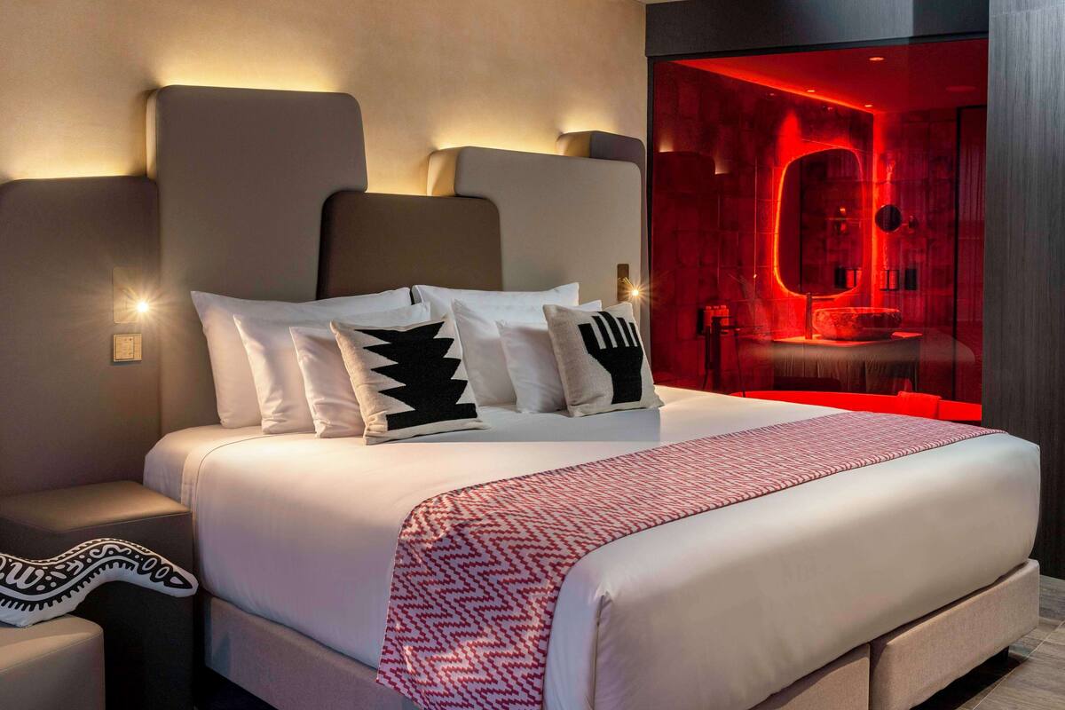 A modern hotel room with a large bed, a nightstand, and a red-lit bathroom visible through a glass partition.