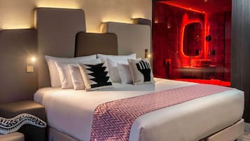 A modern hotel room with a large bed, a nightstand, and a red-lit bathroom visible through a glass partition.