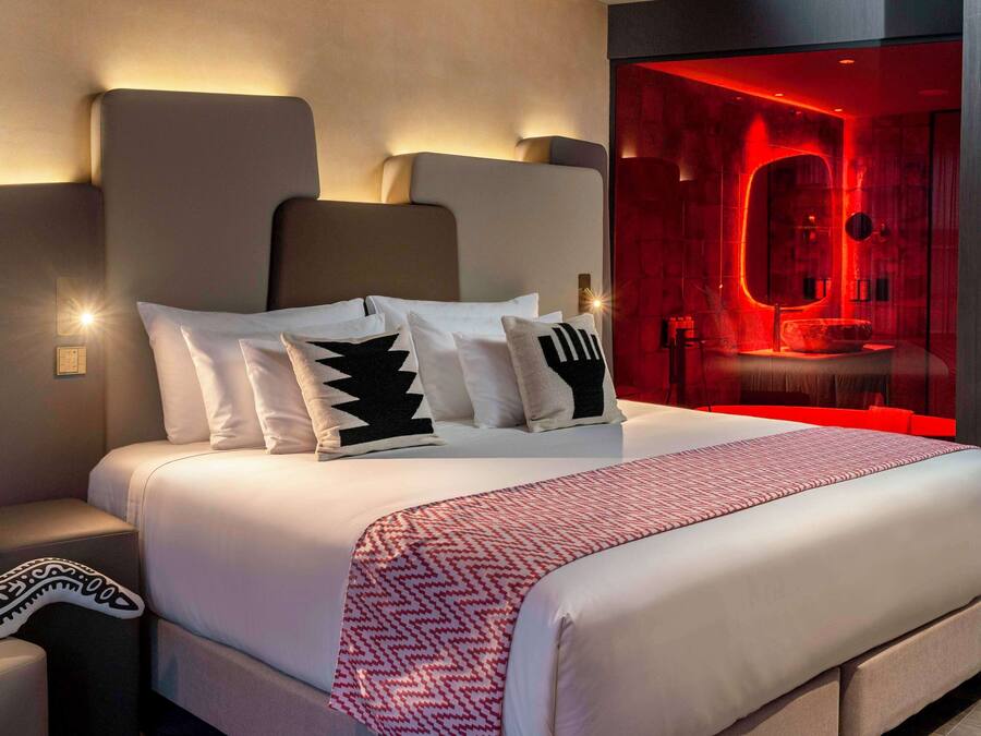 A modern hotel room with a large bed, a nightstand, and a red-lit bathroom visible through a glass partition.