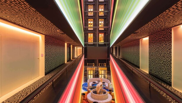 En modern hotellkorridor med väggar som har ett geometriskt mönster, ett centralt, runt sittområde och utsikt över byggnadens interiör.
