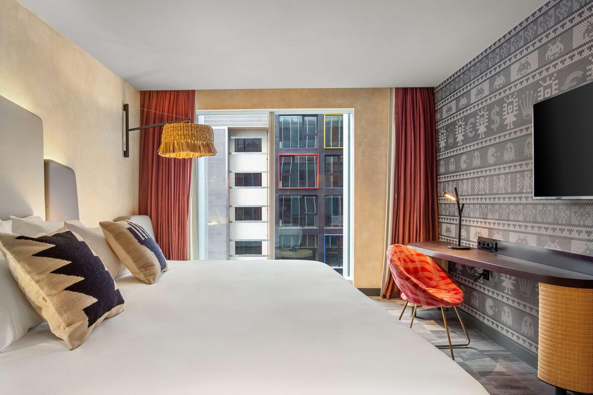 A hotel room with a large bed, a desk, a television, and a view of a building with patterned wallpaper.