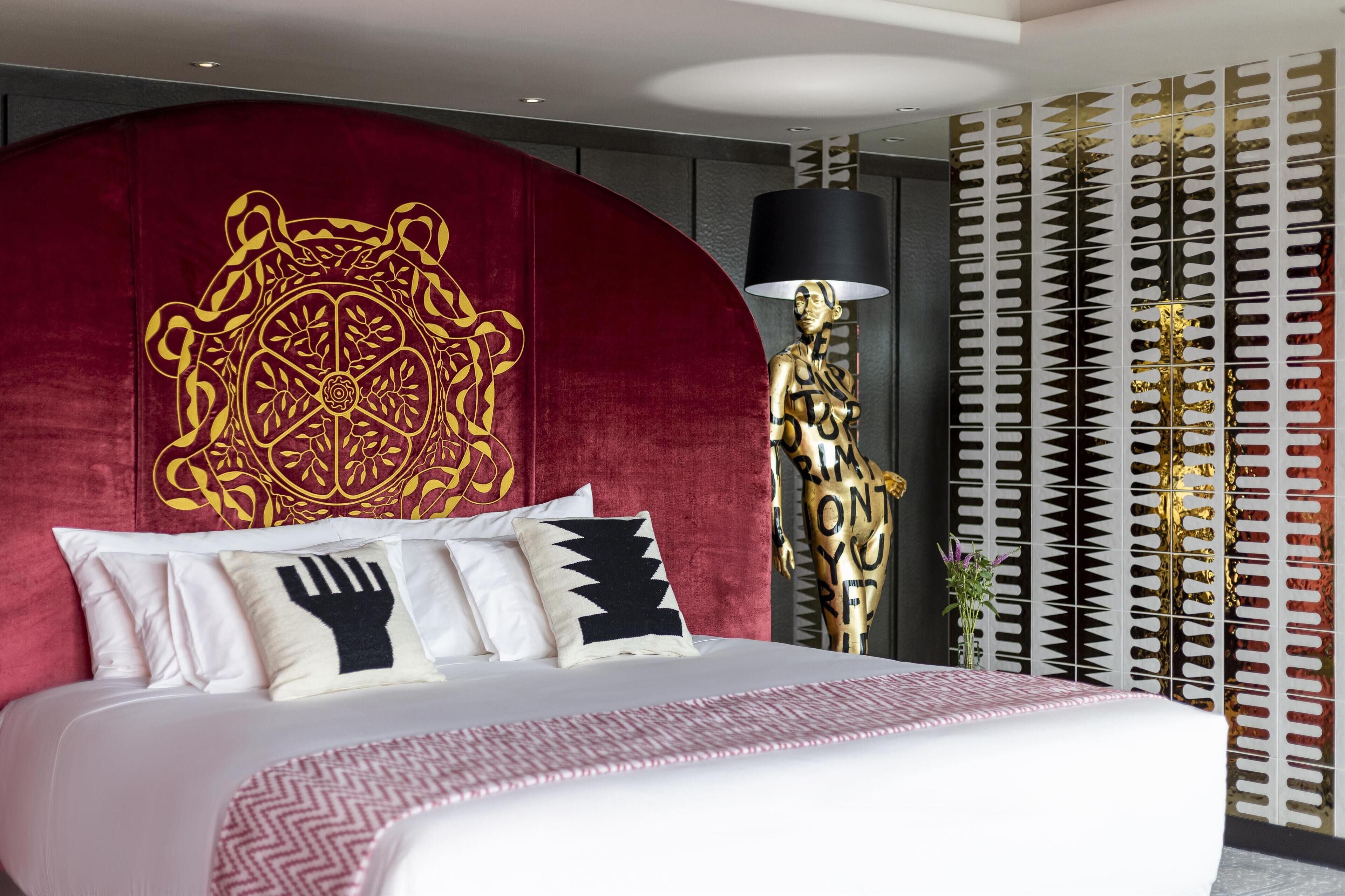 Une chambre d’hôtel moderne avec un grand lit, une tête de lit rouge ornée d’un motif décoratif et un panneau mural à motifs.