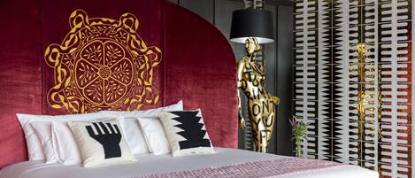 Une chambre d’hôtel moderne avec un grand lit, une tête de lit rouge ornée d’un motif décoratif et un panneau mural à motifs.