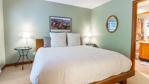 Habitación de hotel con una cama grande, dos mesitas de noche con lámparas, un cuadro en la pared y vistas al baño a través de una puerta abierta.
