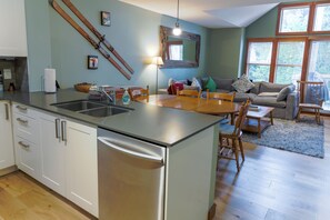 Een moderne keuken met een centraal eiland, een eetgedeelte met een houten tafel en stoelen, en een woonkamer met een bank en een groot raam.