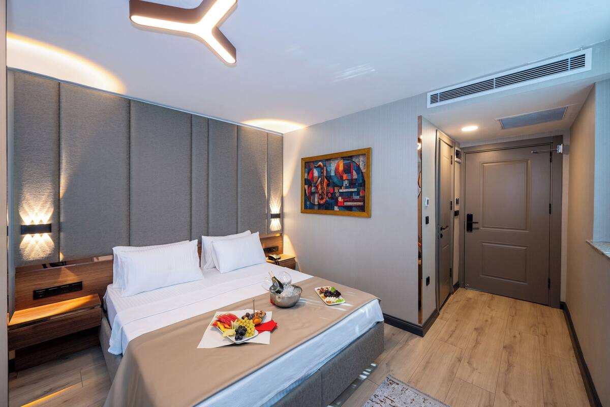 Une chambre d’hôtel moderne avec un lit soigneusement fait, une table de chevet avec une lampe, un tableau au mur et une porte sur le côté droit.