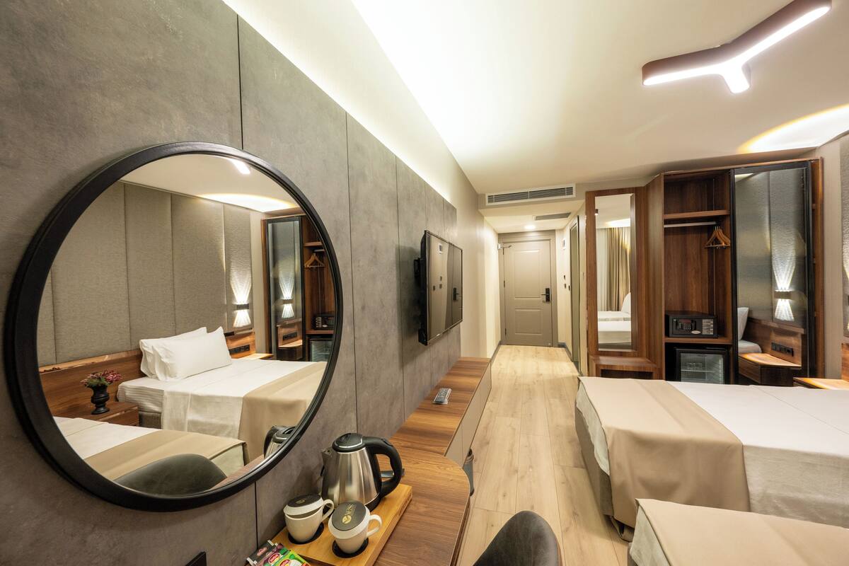 Une chambre d’hôtel avec deux lits, un grand miroir, une table de chevet en bois avec une bouilloire et des tasses, et une armoire.
