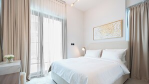 A modern hotel room with a large bed, a bedside table, and a view of the city through sheer curtains. - Trendy 2 Bed in Binghatti Canal (Dubai)